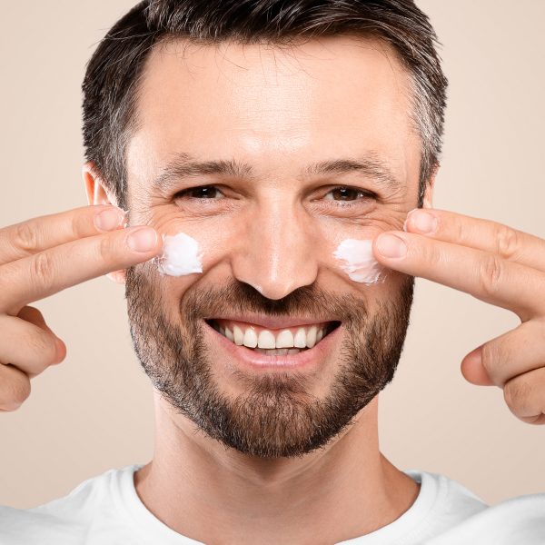 Smiling handsome man applying anti-aging cream on his face, closeup. Middle-aged bearded man with cream on his cheeks from wrinkles and dryness, grey studio background, eye and face care for men