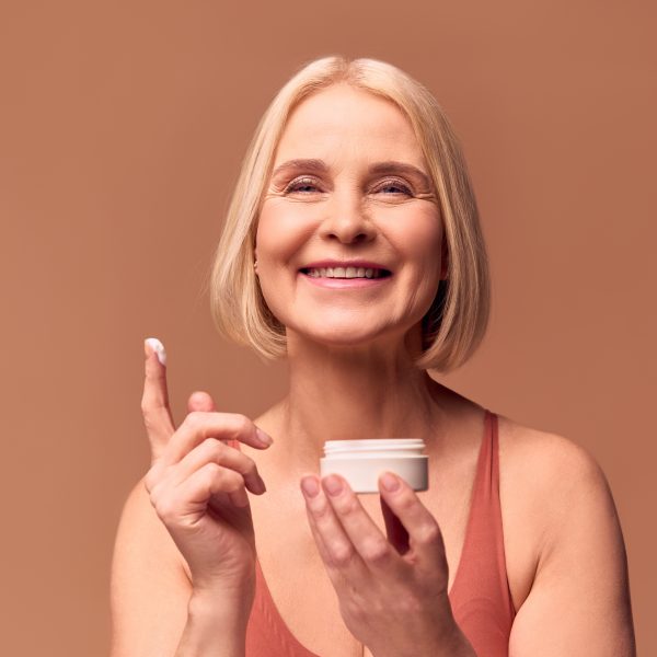 Close-up portrait of a happy beautiful blonde older woman holding a jar of cream in one hand and cream on her finger in the other. Woman laughing and looking at camera on beige background.